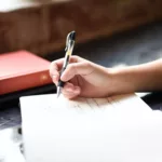 Das Bild zeigt eine Hand mit Stift, die in ein Notizbuch schreibt.